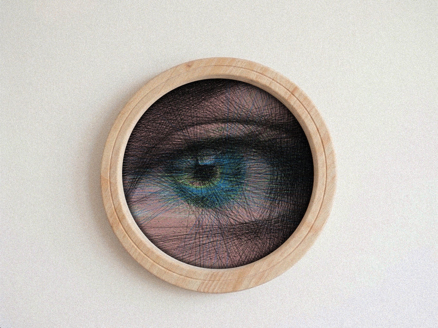 A circular framed string art piece featuring a highly detailed, stylized human eye with blue and green tones, overlaid with black thread, mounted on a light beige wall.