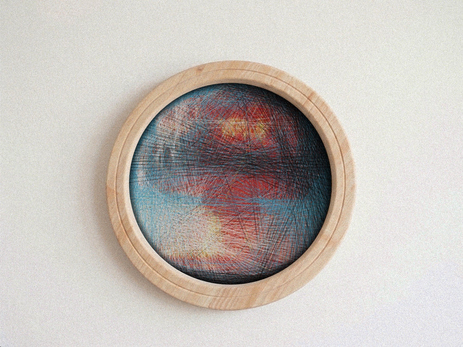 A circular abstract string art piece with red, blue, and yellow lines on a light wood frame, mounted on a textured off-white wall.