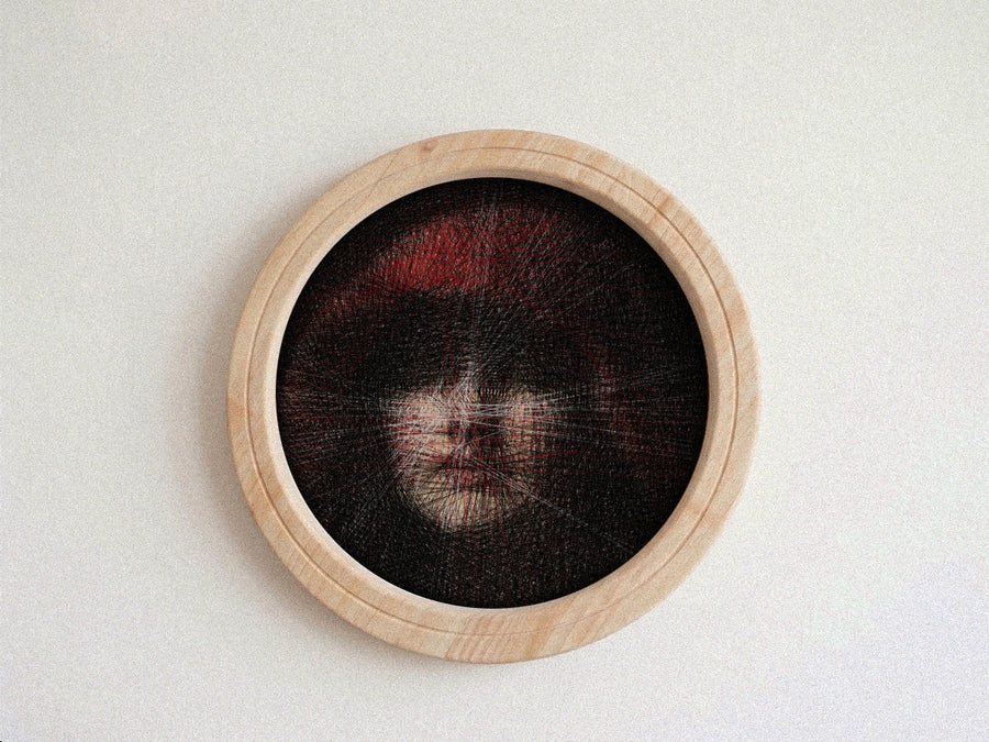 A circular wooden-framed needlework string art piece featuring a highly detailed portrait of a face with dark hair and red hair accents, mounted on a plain light wall.