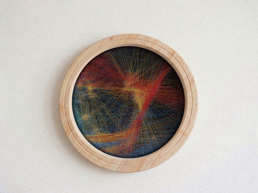 A circular framed abstract string art piece with colorful lines on a dark background, mounted on a light textured wall.