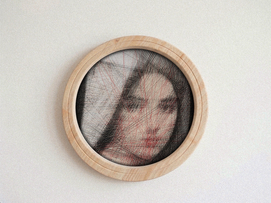 A circular framed portrait string art on a light neutral background. The piece features a partially obscured face with black hair-like lines and red overlay lines, set within a light natural wood frame.