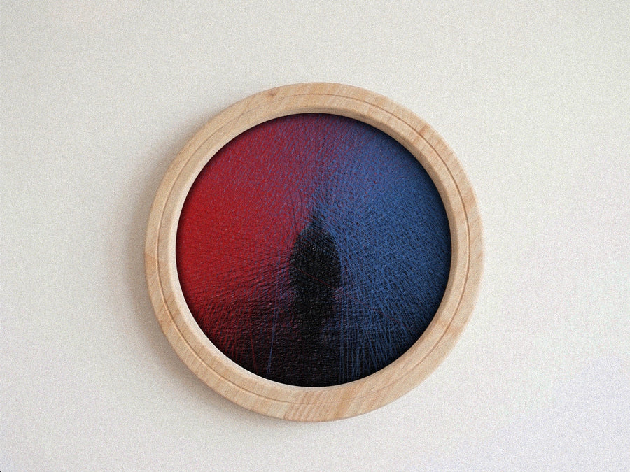 A circular wall string art piece with a light natural wood frame, featuring a textured red-to-blue gradient design with a dark central shape, mounted on a plain off-white wall.