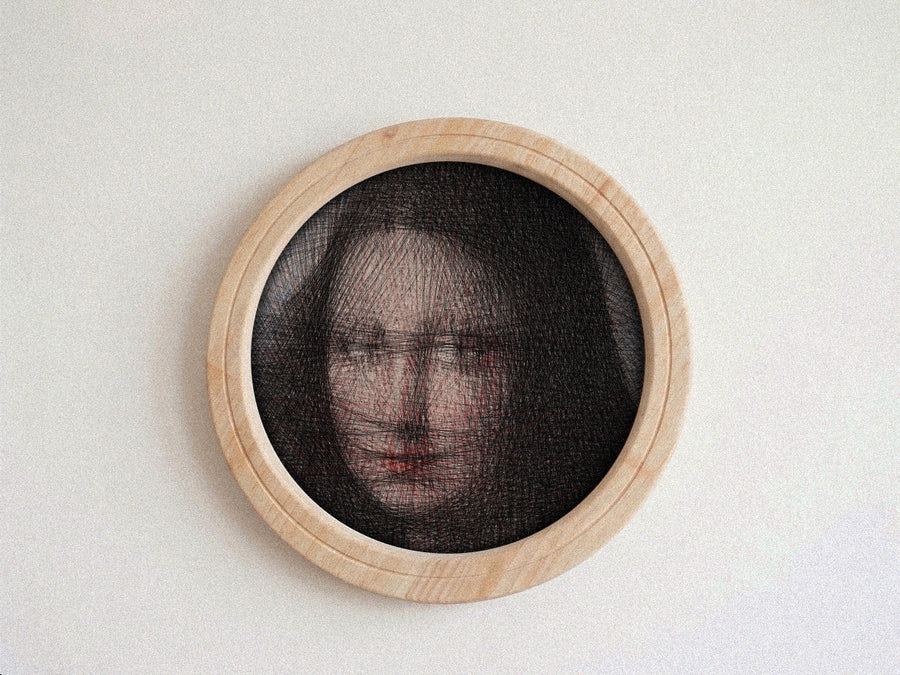 A circular framed string art piece featuring a portrait of a woman's face created with fine black threads over a light wood frame, mounted on a textured off-white wall.