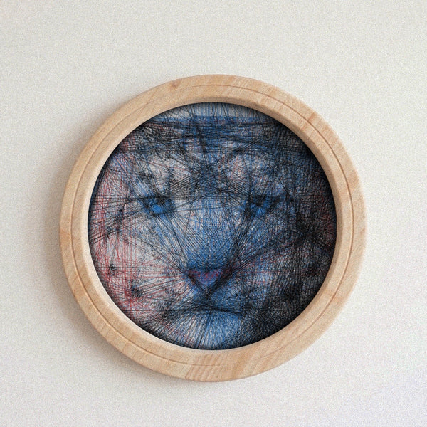 A circular wooden framed string art piece featuring a blue, black, and red abstract string pattern resembling a wolf or cat face, mounted on a light beige wall.