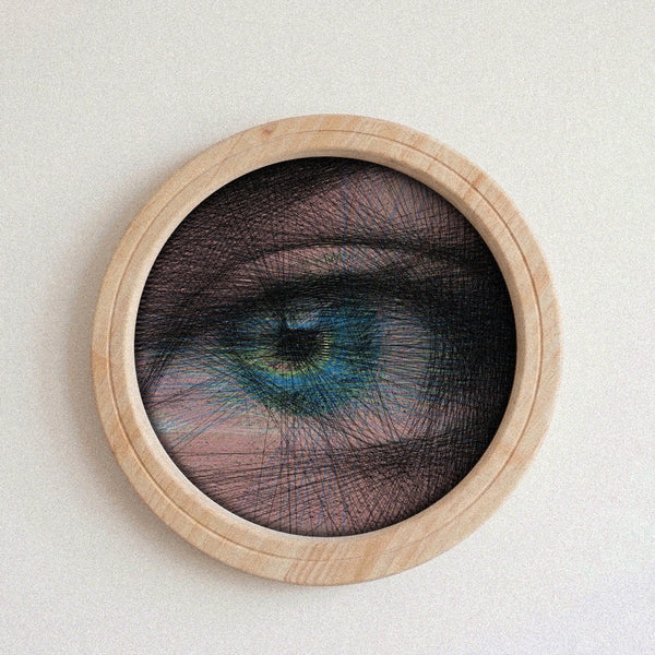 A circular framed string art piece featuring a highly detailed, stylized human eye with blue and green tones, overlaid with black thread, mounted on a light beige wall.