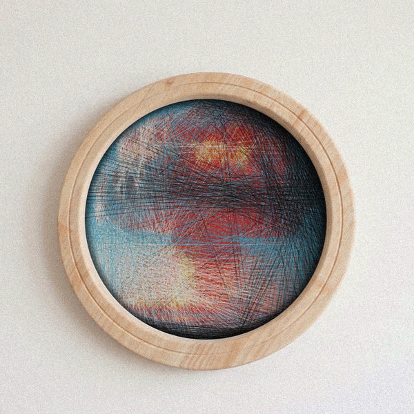 A circular abstract string art piece with red, blue, and yellow lines on a light wood frame, mounted on a textured off-white wall.