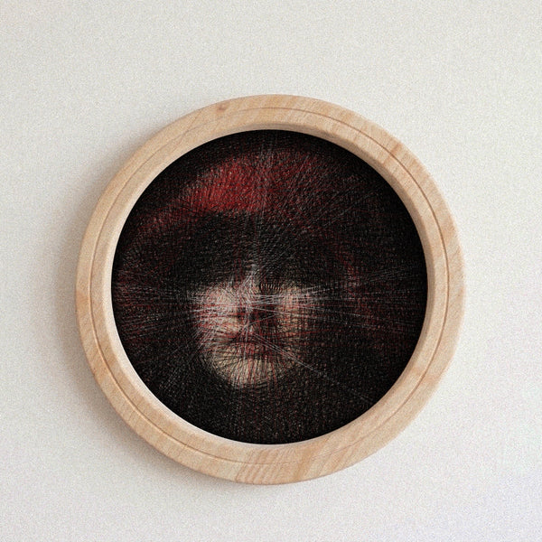 A circular wooden-framed needlework string art piece featuring a highly detailed portrait of a face with dark hair and red hair accents, mounted on a plain light wall.