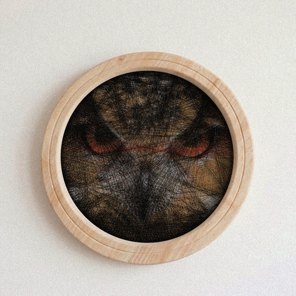 A circular wooden wall string art piece featuring a dark, textured cat face with prominent red eyes, mounted on a plain off-white wall.