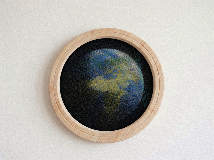 A circular framed string piece featuring a textured, abstract depiction of Earth with blue oceans and green landmasses on a black background, set in a light natural wood frame against a neutral off-white wall.