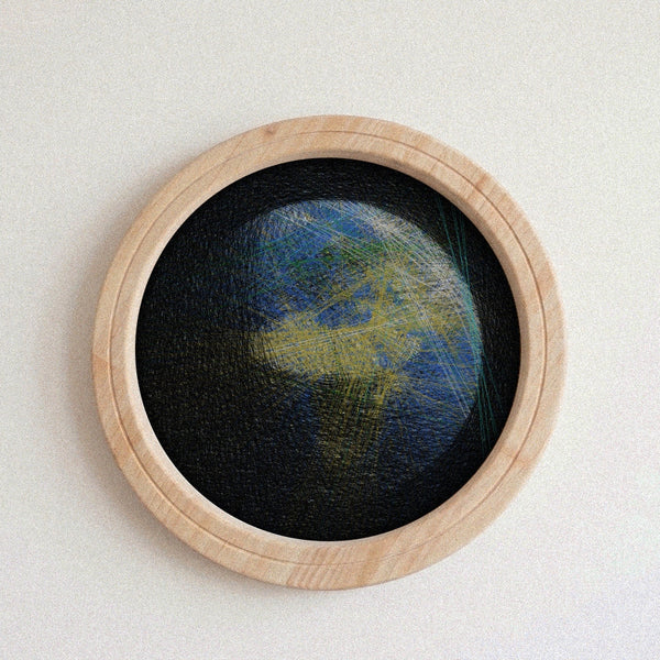 A circular framed string piece featuring a textured, abstract depiction of Earth with blue oceans and green landmasses on a black background, set in a light natural wood frame against a neutral off-white wall.
