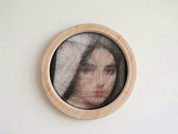 A circular framed portrait string art on a light neutral background. The piece features a partially obscured face with black hair-like lines and red overlay lines, set within a light natural wood frame.