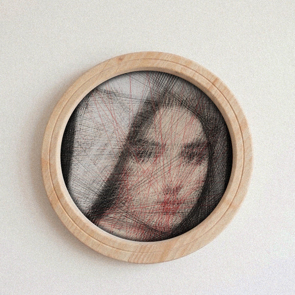 A circular framed portrait string art on a light neutral background. The piece features a partially obscured face with black hair-like lines and red overlay lines, set within a light natural wood frame.