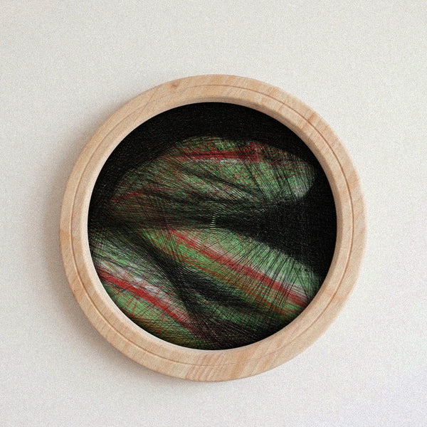 A circular wall string art piece with a light natural wood frame, featuring a black background and a central abstract design made of layered green, red, and black string or thread.