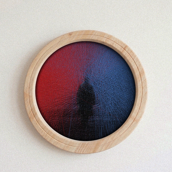 A circular wall string art piece with a light natural wood frame, featuring a textured red-to-blue gradient design with a dark central shape, mounted on a plain off-white wall.