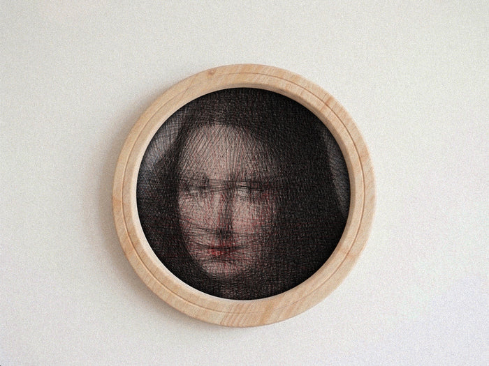 A circular framed string art piece featuring a portrait of a woman's face created with fine black threads over a light wood frame, mounted on a textured off-white wall.