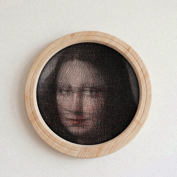 A circular framed string art piece featuring a portrait of a woman's face created with fine black threads over a light wood frame, mounted on a textured off-white wall.